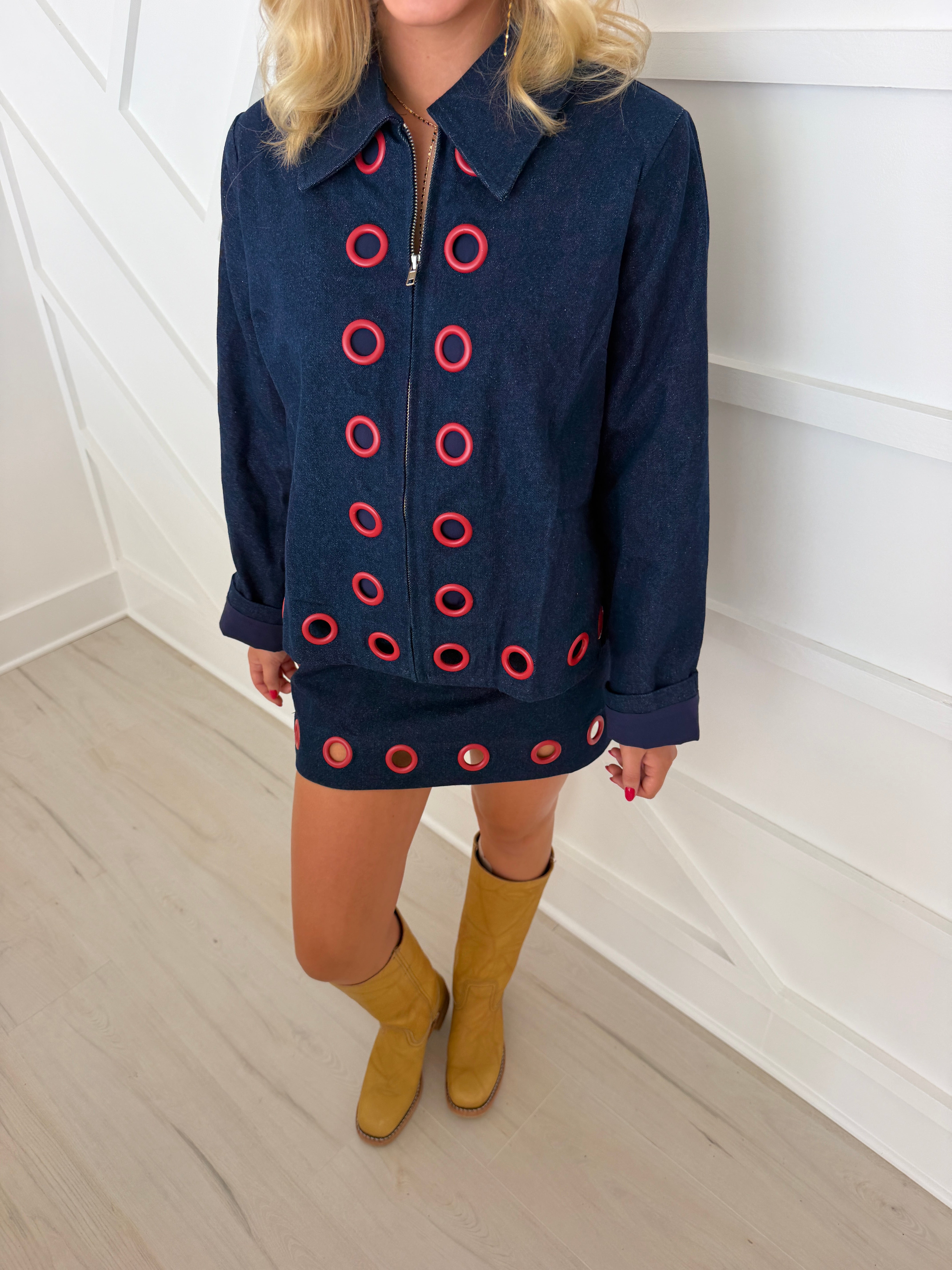 Pretty Follies dark blue denim jacket and mini skirt featuring red eyelets, styled with tan boots on a woman.